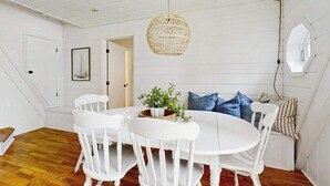 Un intérieur blanc avec une table de salle à manger ronde, des chaises blanches, un luminaire suspendu en fibres tressées et une vue sur l’extérieur par une fenêtre.
