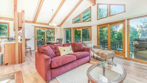 A spacious living area with a red sofa, a glass coffee table, and large windows offering views of the outdoors. - Peaceful, two-story home with a firepit & washer/dryer - close to the beach (Empire)