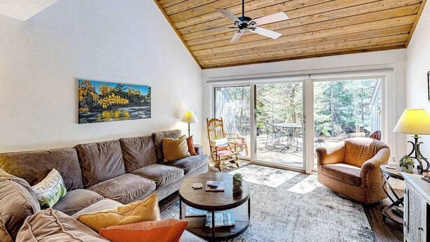 A living room with a wooden ceiling, a sofa, a coffee table, and a painting on the wall. - Riverfront condo with lake access, views, deck, washer/dryer, community firepit (Glen Arbor)