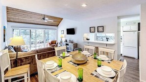 A dining area with a wooden table set for four, surrounded by chairs. There is a sofa and a television in the background. - Riverfront condo with lake access, views, deck, washer/dryer, community firepit (Glen Arbor)