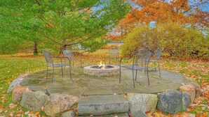 A stone fire pit surrounded by four chairs, set in a stone patio with autumn leaves. - Spacious, airy home near Little Glen Lake with gas grill, deck, and new firepit (Empire)