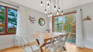 A dining area with a wooden table and white chairs, large windows with curtains, and a wall-mounted light fixture. - Spacious, airy home near Little Glen Lake with gas grill, deck, and new firepit (Empire)