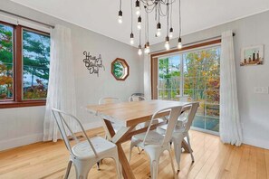 Ein Essbereich mit Holztisch und weißen Stühlen, großen Fenstern mit Vorhängen und einer Wandleuchte.