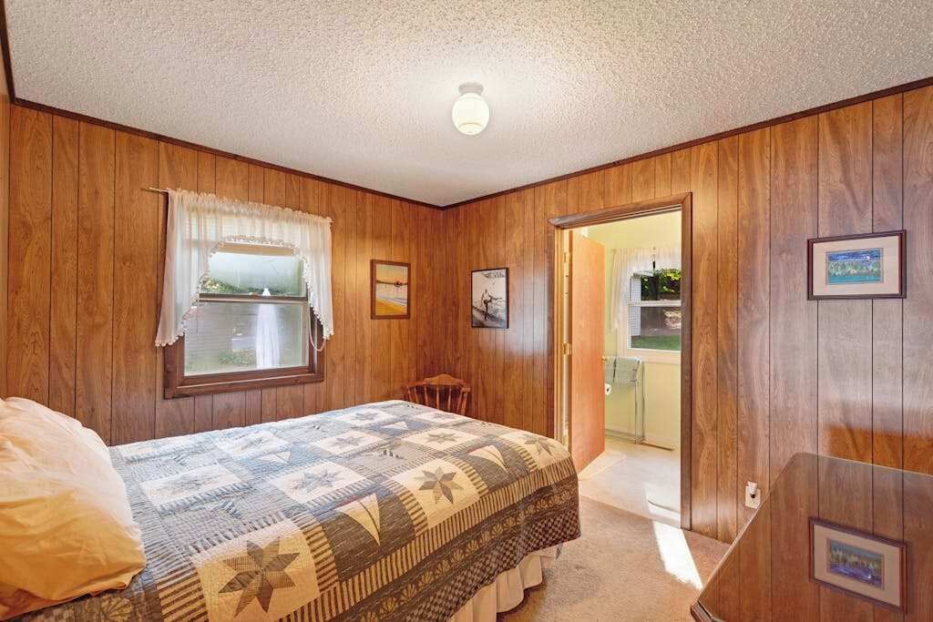 A bedroom with wooden walls, a bed with a patterned bedspread, a window with curtains, a small table, and a door leading to another room.