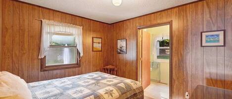 A bedroom with wooden walls, a bed with a patterned bedspread, a window with curtains, a small table, and a door leading to another room.