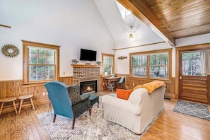 A cozy living room with a fireplace, wooden floors, and large windows.