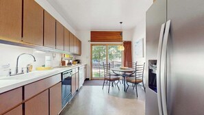 A kitchen with wooden cabinets, a stainless steel refrigerator, a dining table with chairs, and a window with a view of greenery. - Rustic beach cottage with partial lake views, grill, & WiFi - great location (Michiana)