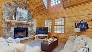 Ein gemütliches Blockhäuschen-Interieur mit Kamin aus Stein, einem Fernseher und einem großen Fenster.