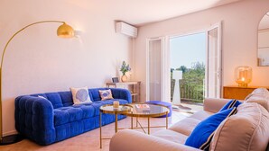 A living room with a blue sofa, a beige sofa, a round glass coffee table, a floor lamp, and a sliding glass door leading to a balcony. - Le Pioulier – Villa in Vence with an open view (Vence)
