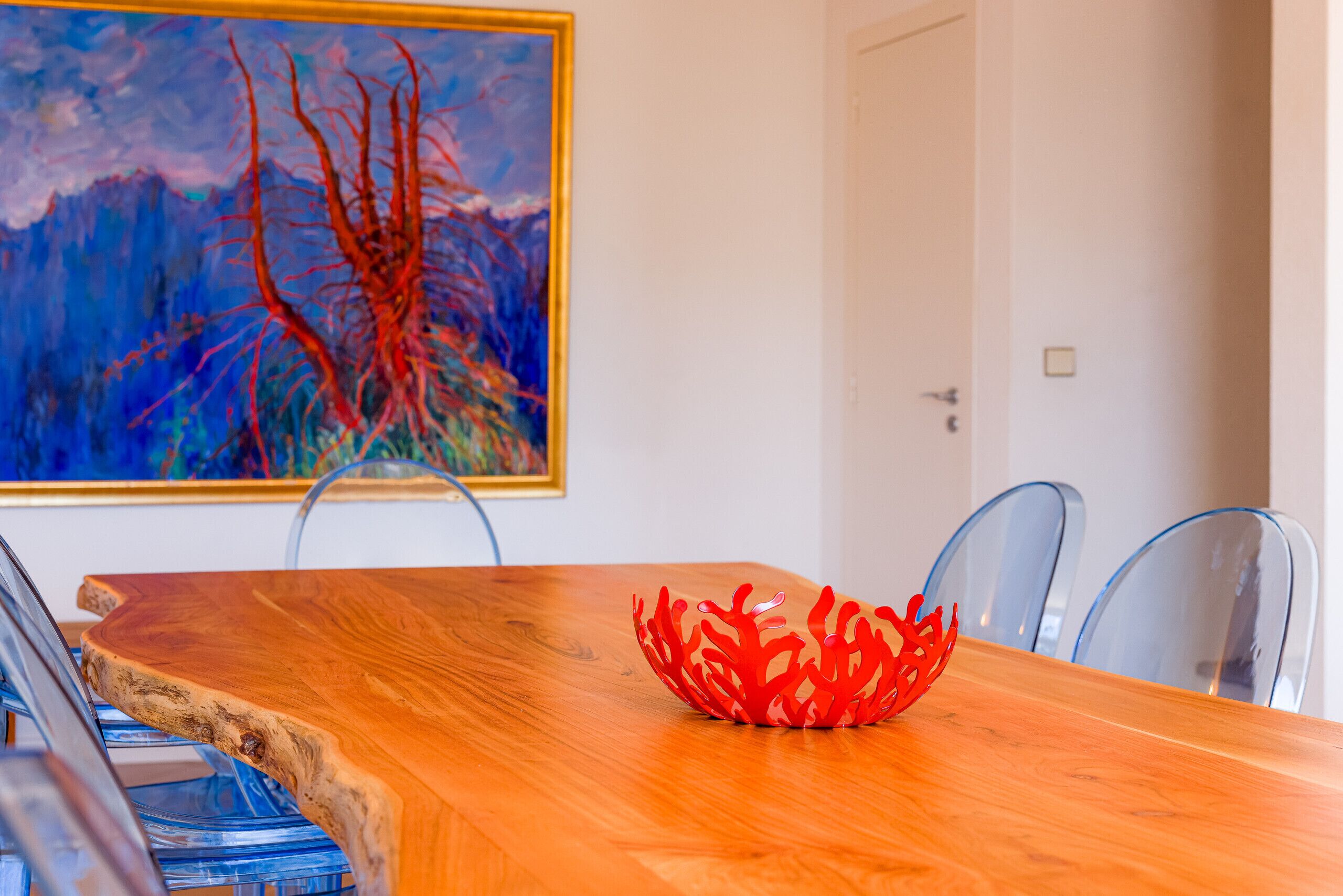 Een houten eettafel met een rode, koraalachtige decoratie in het midden, transparante glazen stoelen en een schilderij aan de muur.