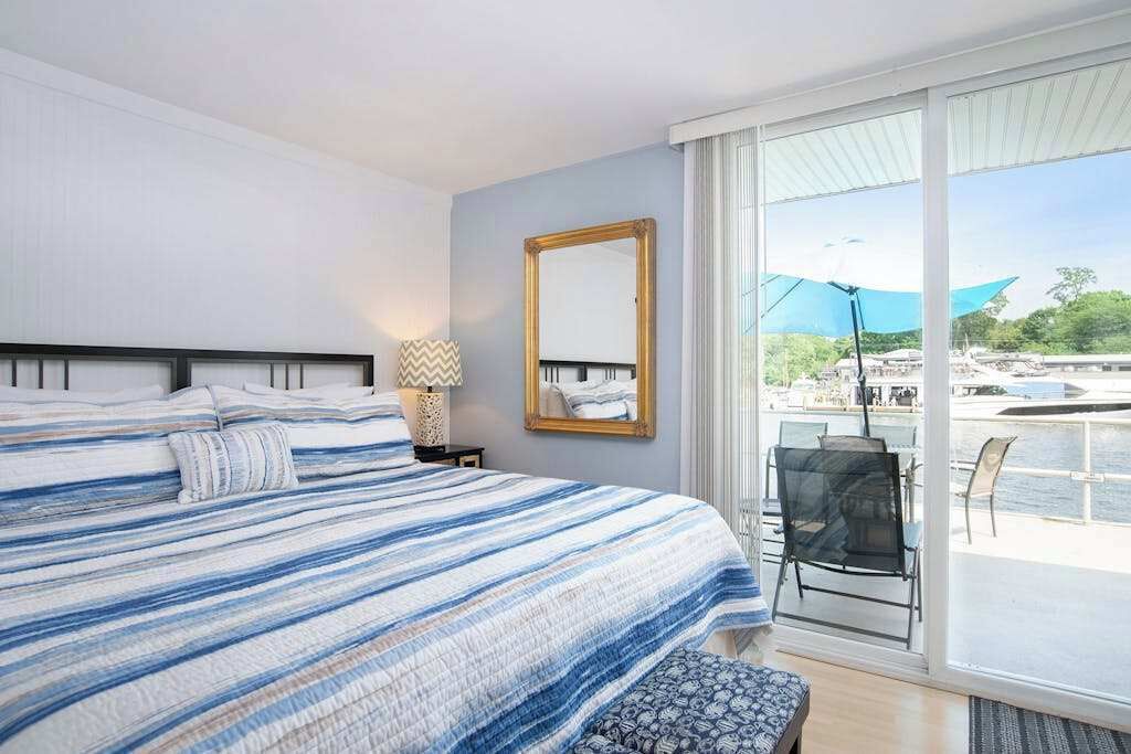 A bedroom with a large bed, a bedside table with a lamp, a mirror, and a view of the outside.