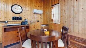 A wooden-paneled room with a round wooden table, four chairs, a clock, an air conditioning unit, and a window with curtains. - Peaceful wooded escape w/ kitchenette, gas grill, WIFI, lakes & trails nearby (Covert)