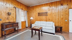 A wooden-paneled room with a white sofa, a small table, a lamp, and a vintage telephone. - Peaceful wooded escape w/ kitchenette, gas grill, WIFI, lakes & trails nearby (Covert)