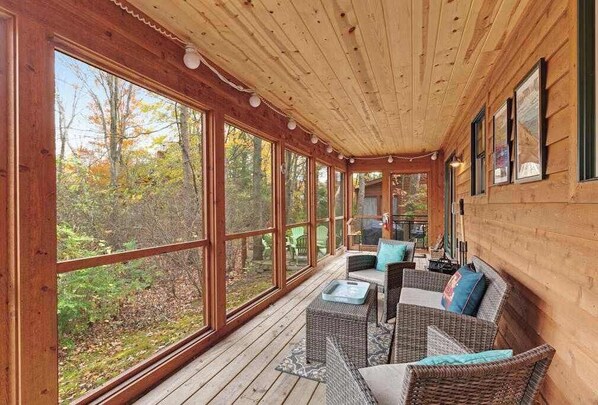 A covered porch with large windows, wicker furniture, and a view of the forest.