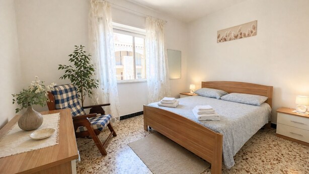 A bedroom with a bed, bedside tables, a chair, a desk, a potted plant, and a window with curtains.