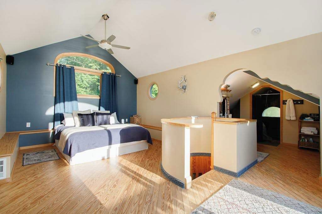 Une chambre avec un lit, un ventilateur de plafond, une fenêtre avec des rideaux, une petite salle de bain avec un lavabo et un escalier.