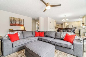 A modern living room with a grey sectional sofa, red and patterned cushions, a ceiling fan, and a kitchen in the background.