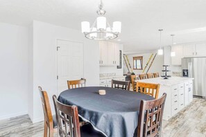 A dining area with a round table, chairs, a chandelier, and a kitchen in the background.