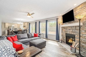 A modern living room with a fireplace, a flat-screen TV, and a dining area in the background.