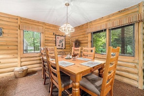A wooden cabin interior with a dining table, chairs, a chandelier, and large windows.