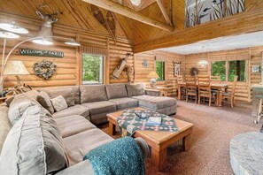 Interior de una cabaña con un sofá modular, una mesa de comedor de madera con sillas, una mesa de centro acolchada y un letrero de bienvenida.