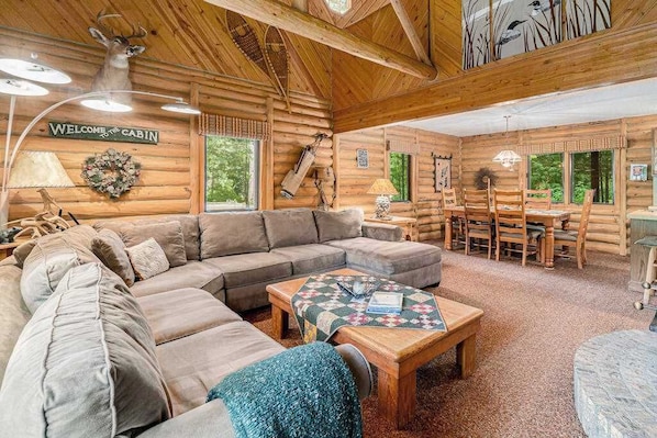 A cabin interior with a sectional sofa, a wooden dining table with chairs, a quilted coffee table, and a welcome sign.