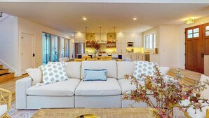 A modern living room with a sofa, a coffee table, and a kitchen in the background. - Brand New Luxurious Home, w/ Fire pit, Gas Grill, near Lakes & Trails! (Glen Arbor)