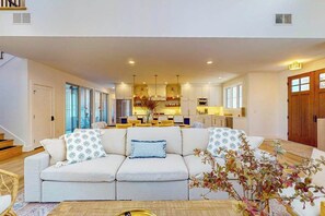A modern living room with a sofa, a coffee table, and a kitchen in the background. - Brand New Luxurious Home, w/ Fire pit, Gas Grill, near Lakes & Trails! (Glen Arbor)