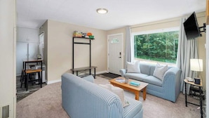 A living room with a sofa, a coffee table, a TV, and a window with a view of greenery. - Private dog-friendly hideaway w/  firepit - close to Crystal Mountain (Thompsonville)