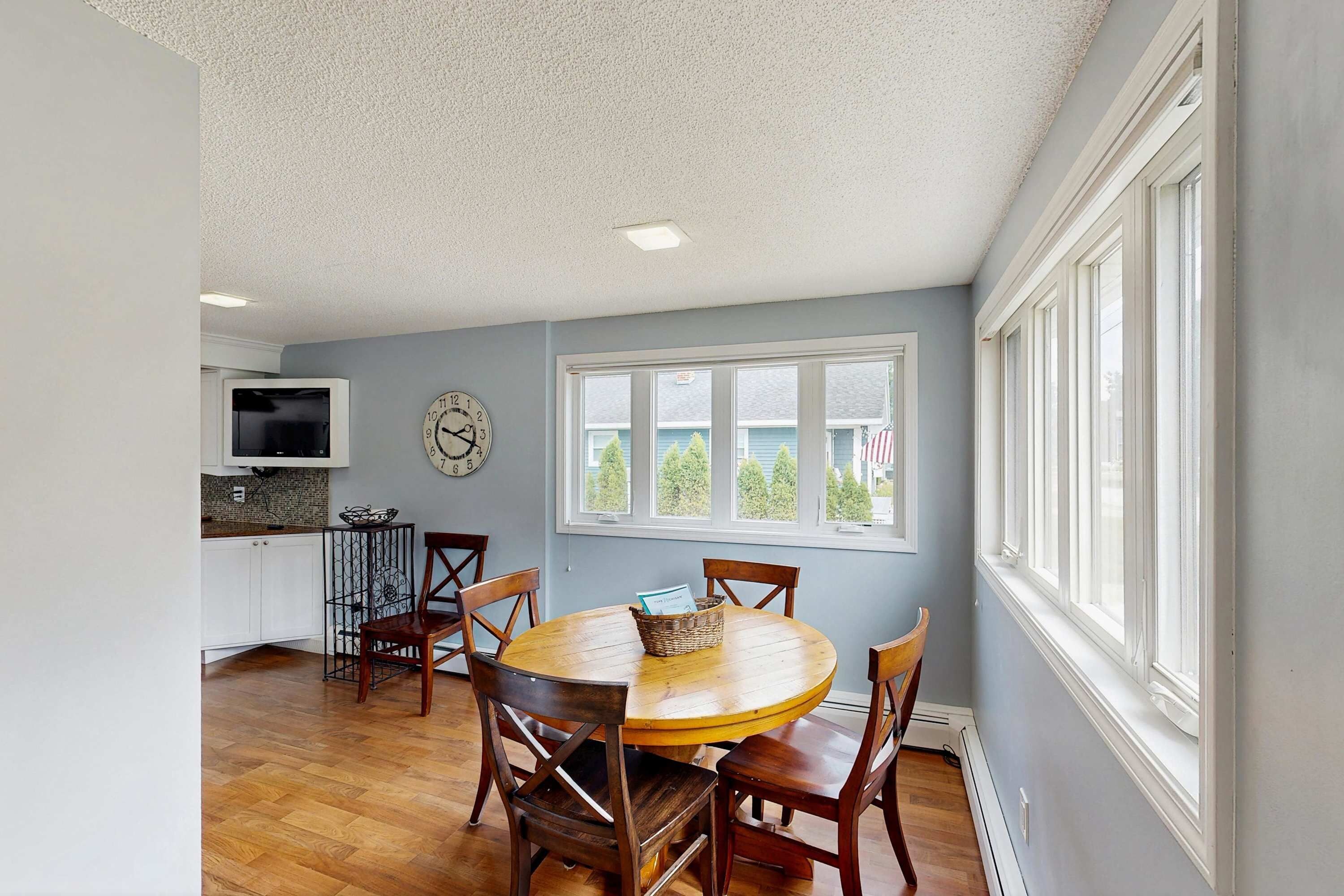 A dining area with a round wooden table and chairs, a kitchen with white cabinets and a TV, and a large window with a view of trees.