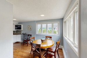 A dining area with a round wooden table and chairs, a kitchen with white cabinets and a TV, and a large window with a view of trees. - Dreamy Coastal Cottage W/ Free WiFi, Full Kitchen, & Large Deck - Walk to Town (Harbor Springs)