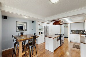 A modern kitchen with white cabinetry, stainless steel appliances, and a wooden dining table with bar stools. - Dreamy Coastal Cottage W/ Free Wi-Fi, Full Kitchen, & Large Deck - Walk to Town (Harbor Springs)
