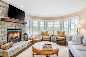 A living room with a stone fireplace, a round leather ottoman, two plaid chairs, a sofa, and a TV mounted above the fireplace.