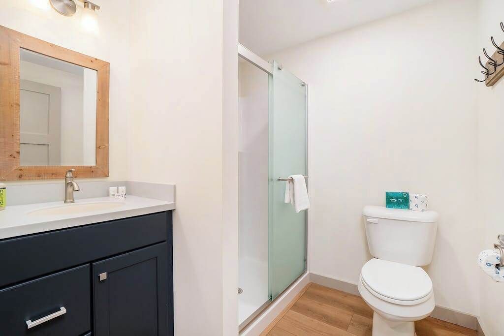 Un baño con un espejo de marco de madera, un lavamanos blanco, un mueble negro, una cabina de ducha con paredes de vidrio y un inodoro blanco.