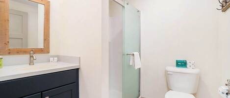 Un baño con un espejo de marco de madera, un lavamanos blanco, un mueble negro, una cabina de ducha con paredes de vidrio y un inodoro blanco.
