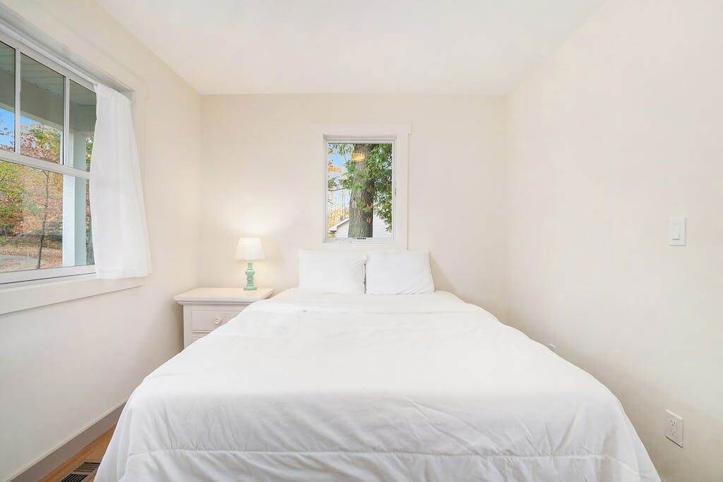 A bedroom with a bed, a nightstand, a lamp, a window with curtains, and a view of trees outside.