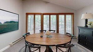 A dining room with a round wooden table, six chairs, a vase with flowers, and a view through large wooden-framed windows. - Single-level condo with a full kitchen, & washer/dryer (Glen Arbor)