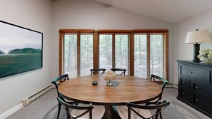Une salle à manger avec une table ronde en bois, six chaises, un vase rempli de fleurs et une vue dégagée sur l’extérieur grâce à de grandes fenêtres à cadre en bois.
