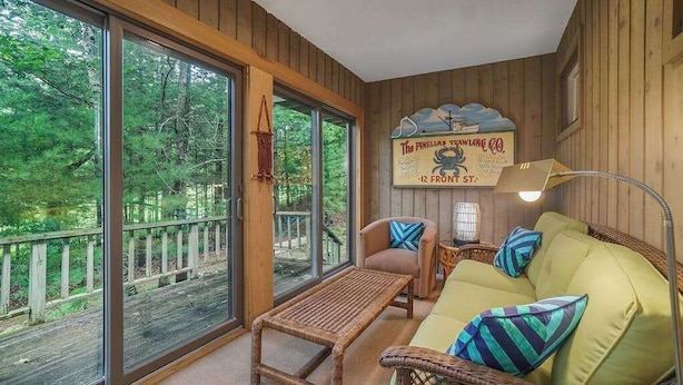 A cozy cabin interior with a large sliding glass door leading to a deck, a wicker sofa with cushions, a small table, and a sign on the wall. - Single-level condo with a full kitchen, & washer/dryer (Glen Arbor)