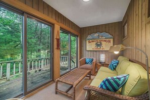 L’intérieur chaleureux d’un chalet, avec une grande porte-fenêtre coulissante donnant sur une terrasse, un canapé en osier avec des coussins, une petite table et un panneau accroché au mur.