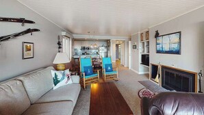 A living room with a sofa, a coffee table, and a fireplace. There is a dining area with chairs and a table. - Tasteful Glen Arbor condo with beautiful Lake Michigan views, deck & fireplaces (Glen Arbor)
