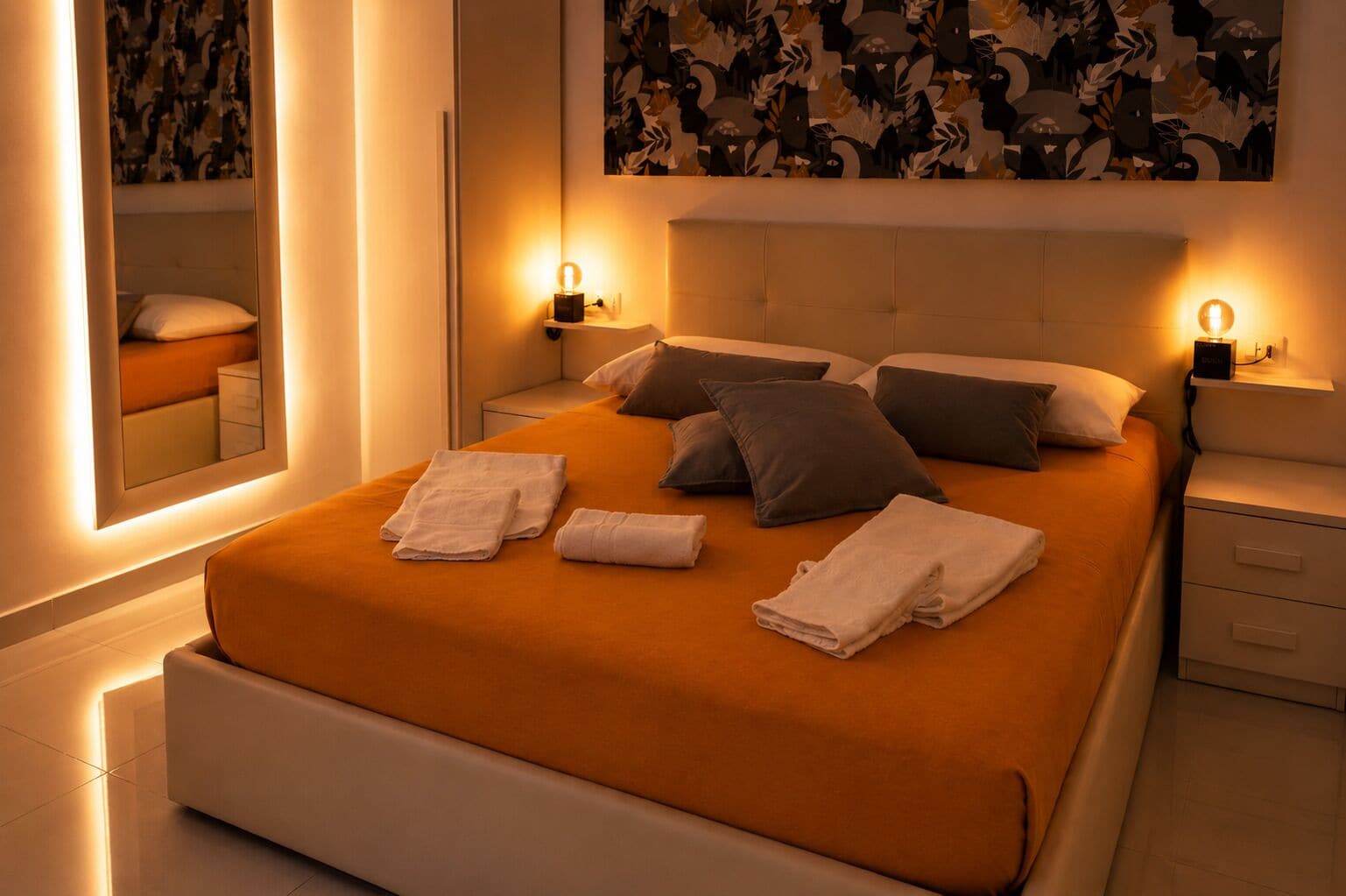 A neatly made bed with pillows and towels, bedside tables with lamps, a large mirror, and a wall with abstract art.