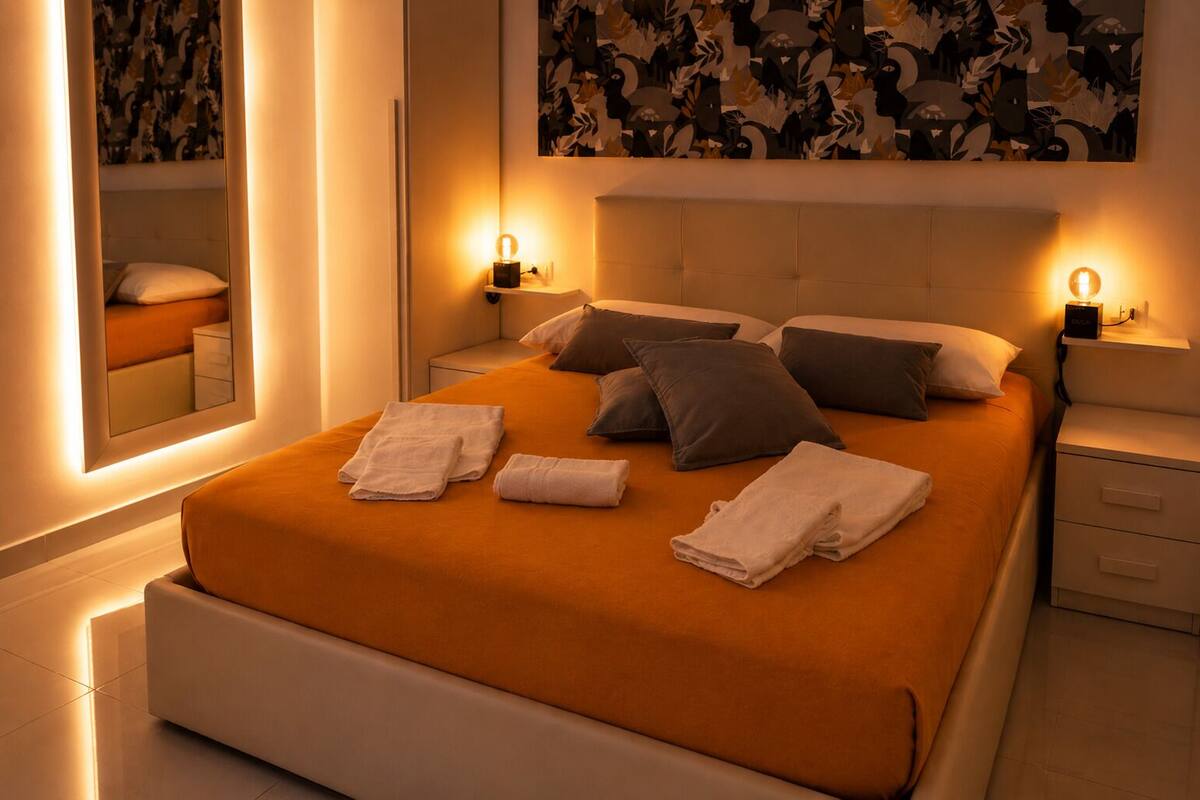 Cama bem arrumada com travesseiros e toalhas, mesas de cabeceira com abajures, um espelho grande e uma parede com arte abstrata.