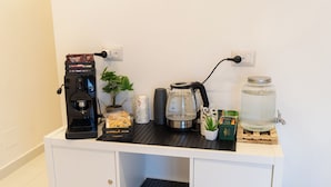 Una kitchenette con cafetera, hervidor, un tarro y una pequeña planta sobre un mueble blanco.