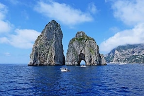 Two large rock formations with a natural archway, a boat in the water, and a mountainous coastline in the background. - Vesun Suites (Naples)