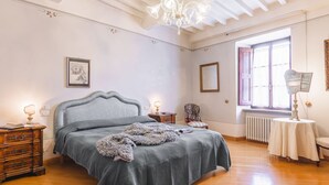 A bedroom with a bed, wooden dresser, nightstands, a chair, a table, a chandelier, a window, and a radiator. - Palazzo Simonetti - a noble elegance in the heart of Cortona (Cortona)