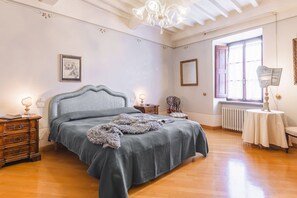 A bedroom with a bed, wooden dresser, nightstands, a chair, a table, a chandelier, a window, and a radiator. - Palazzo Simonetti - a noble elegance in the heart of Cortona (Cortona)