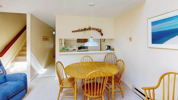 Een eetruimte met een ronde houten tafel en zes stoelen, een keuken met een wit aanrecht en apparatuur, en een trap naar een hoger niveau.