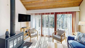 Een gezellige woonkamer met een open haard, houten plafond, grote ramen en een flatscreen televisie.