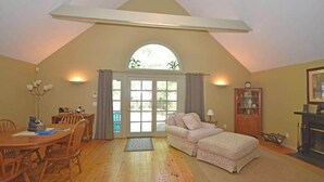 A room with a dining table, two chairs, a sofa, a fireplace, and a large arched window. - Charming home near the lake with a fireplace, central AC, washer/dryer & grill (Glen Arbor)